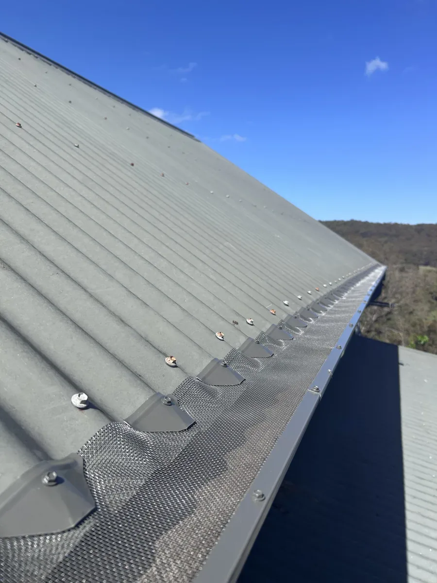 Metal ember mesh held by screwed saddle clips along the roof crest and a screw-fixed edging strip over the gutter, with bushland hills behind
