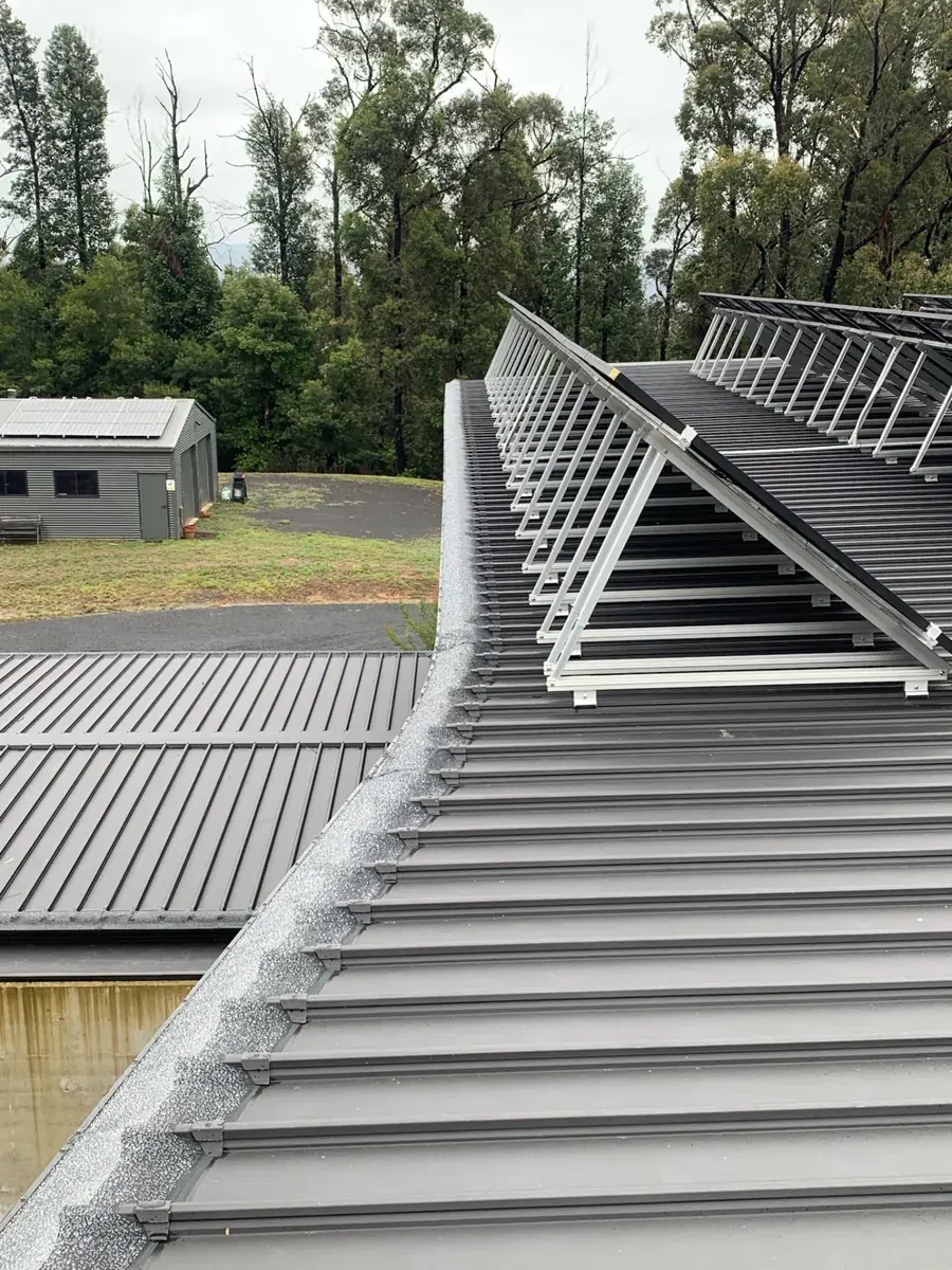 Metal ember mesh screwed to trim strips along a curved dark metal roofline on a Kangaroo Valley home, with solar panels above and eucalypts behind
